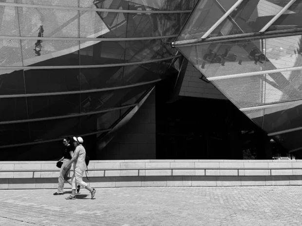 The Fondation Louis Vuitton in black and white: a black glass sail filling the frame with two small figures walking past the foot of it.