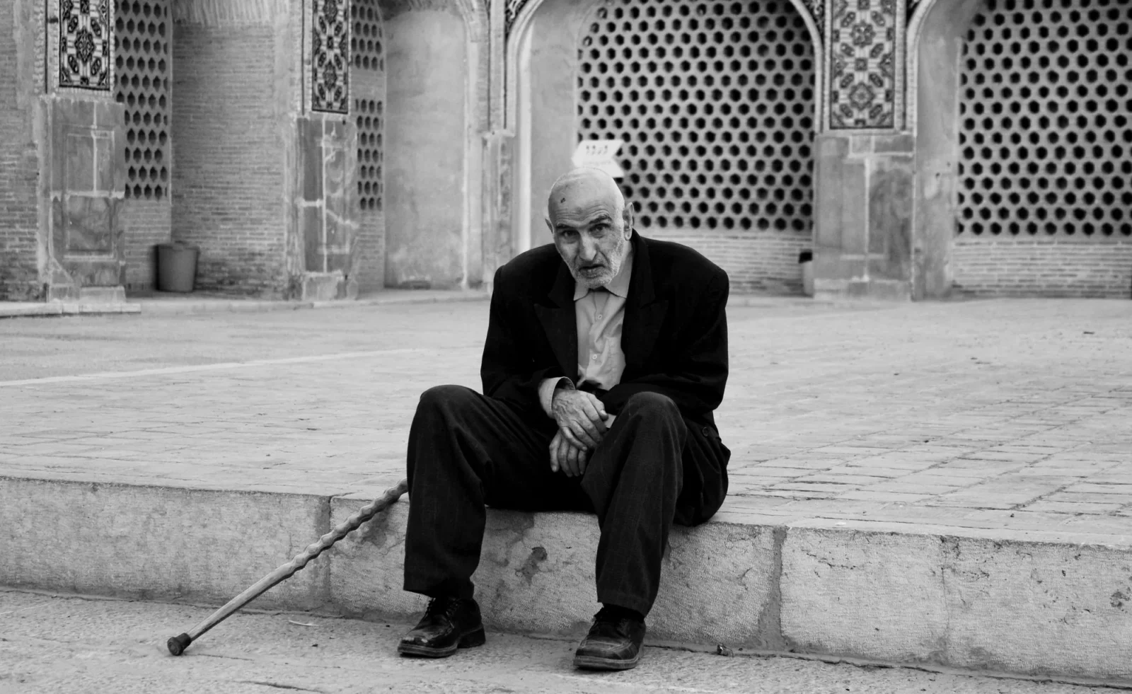 A man with a cane on a mosque step, tilework behind, black and white.