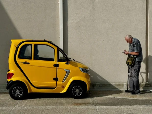 A yellow micro-car and an old man with a stick against a blank wall, both lit the same way.