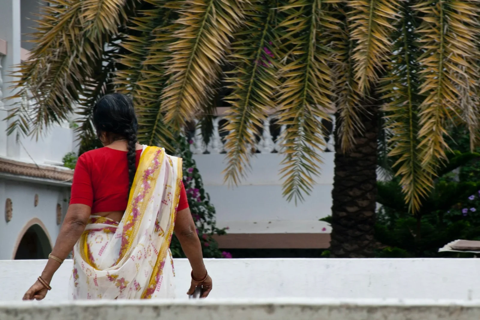 A woman in a red and yellow sari, back turned, under palm fronds.