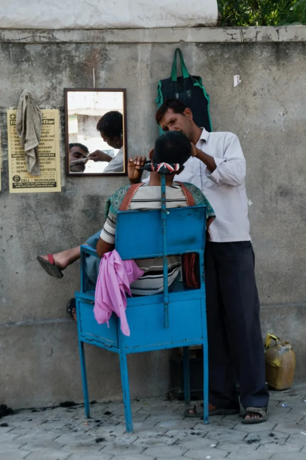 A street barber at a blue chair, a mirror hung on the concrete behind him.