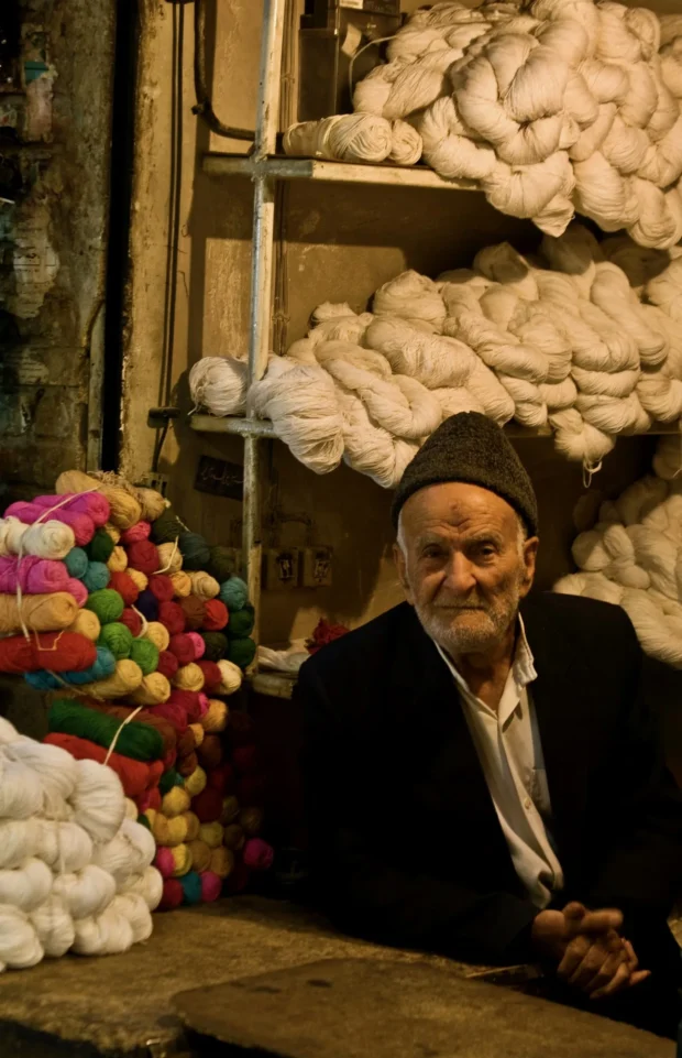 The same seller alone, facing the camera, the dyed wool stacked to the ceiling around him.