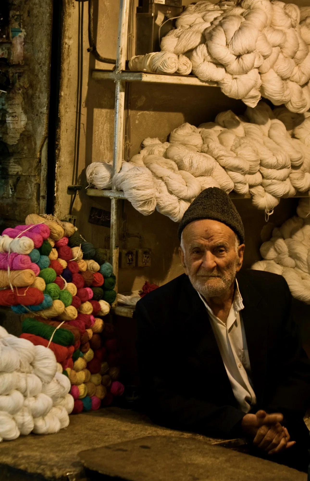 The same seller alone, facing the camera, the dyed wool stacked to the ceiling around him.